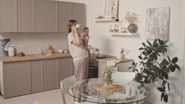 Medium long of young Biracial woman in loungewear holding adorable infant, standing in kitchen at daytime, drinking coffee from mug