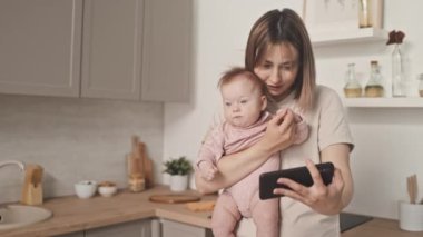Medium of brown-haired young Biracial woman holding cute infant in pink clothing, standing in kitchen at daytime, video calling via smartphone