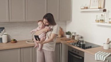 Orta boylu, kahverengi saçlı, melez bir kadının kucağında tatlı bir kız çocuğu, gündüz vakti mutfakta dikilip, akıllı telefondan video görüşmesi yapması.