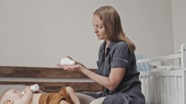 Tilting down of young Caucasian woman sitting on bed at home in afternoon, applying moisturizer on legs of cute blond-haired toddler boy