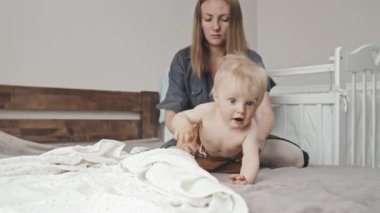 Low angle of young Caucasian woman sitting on bed at home in afternoon, dressing cute blond-haired toddler boy