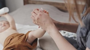 Over-shoulder of cropped parent sitting on bed at home in afternoon, applying moisturizer on feet of blurred toddler boy