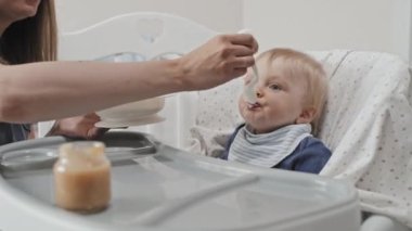 Low angle of cropped mother feeding with childrens food cute blue-eyed Caucasian toddler boy who sitting high chair at daytime