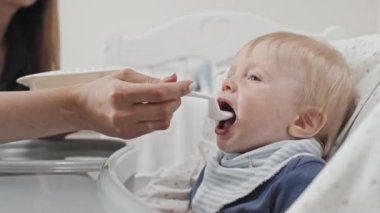 Low angle of unrecognizable caregiver feeding with childrens food cute blue-eyed Caucasian toddler boy who sitting high chair at daytime