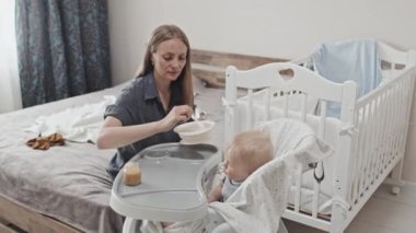 High angle of young beautiful Caucasian woman feeding with childrens food blond-haired Caucasian toddler boy who sitting high chair at daytime in nursery