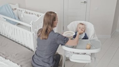 High angle of young female Caucasian babysitter feeding with childrens food blue-eyed Caucasian toddler boy who sitting high chair at daytime in nursery