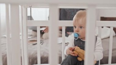 Medium long of adorable blond-haired Caucasian boy sucking pacifier, sitting in crib, playing with toys at daytime