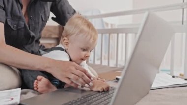 Low angle of young Caucasian mother sitting on bed with her cute toddler son, working with documents and portable computer, talking on cellphone, boy playing with device