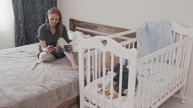 Wide shot of young Caucasian mother sitting on bed in bedroom at daytime, drinking coffee, scrolling social media feed on smartphone, her toddler son playing in crib