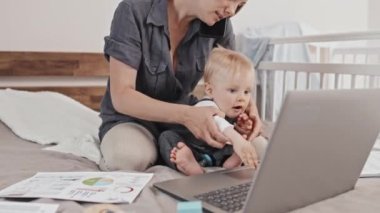 Tilting up of young Caucasian mother sitting on bed with her cute toddler son, working with documents and portable computer, talking on cellphone