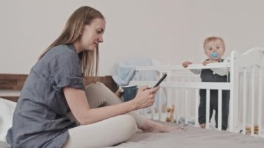 Tracking in of young Caucasian mother sitting on bed in bedroom at daytime, drinking coffee, scrolling social media feed on smartphone, her toddler son standing in crib