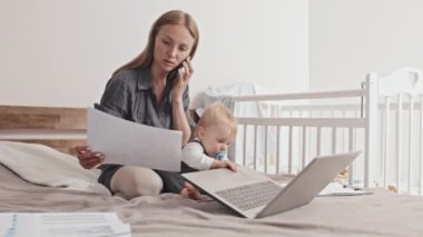 Full shot of young Caucasian woman sitting on bed with her toddler son, working with documents and portable computer, talking on mobile phone