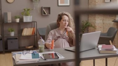 Medium of young long-haired Caucasian woman sitting at desk in workplace at daytime, snacking with fresh sliced veggies, using portable computer