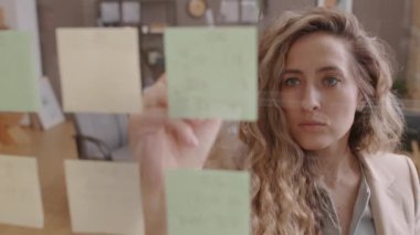 Medium of young blonde blue-eyed Caucasian businesswoman standing in office, writing on sticky notes on glass wall in afternoon