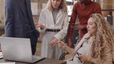 Tilting up of long-haired expecting woman sitting at desk, Black man and Caucasian female and male colleagues standing on her side, talking and looking at laptop computer