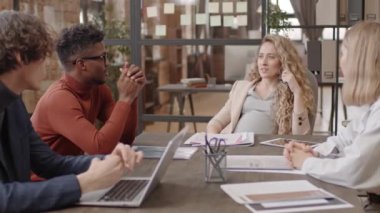 Medium of pregnant woman, African American man and Caucasian female and male colleagues sitting at conference table, having business meeting and talking in afternoon