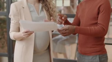 Midsection of blonde cropped expectant businesswoman holding documents, talking to unrecognizable male employee, standing in office at daytime