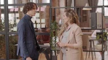 Side view of curly pregnant manager in formalwear talking to young male Caucasian colleague, standing in workplace at daytime