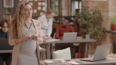 Portrait of curly blond-haired Caucasian mother-to-be in formalwear standing in modern office at daytime, looking and smiling on camera