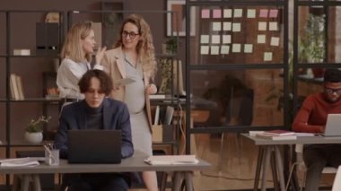 Tracking of blond-haired Caucasian mother-to-be and her young girlfriend walking between desks in coworking space, chatting, smiling