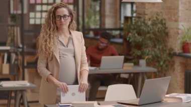 Portrait of curly blond-haired Caucasian mother-to-be in eyeglasses and formalwear standing in modern office at daytime, looking and smiling on camera