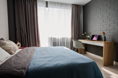 Double bed with pastel cushions and blue duvet standing by wall in the center of spacious cozy bedroom with workplace of freelancer