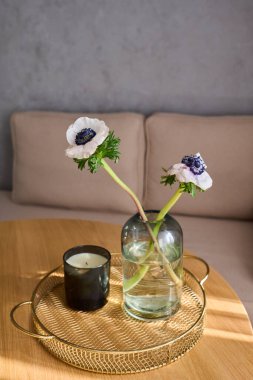 Two wildflowers with long stems standing in jar with water next to scented candle in black glass in round handmade wicker tray on table