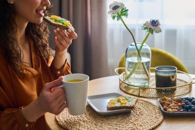 Side view of young hungry female vegetarian having sandwich with cheese, ruccola and tomatoes with fresh orange juice
