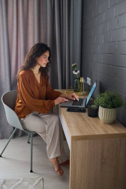 Serious female freelancer typing on laptop keyboard while sitting by wooden desk in front of grey brick wall and working from home