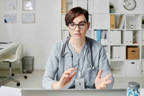 Confident doctor in medical scrubs communicating to online patients while sitting by desk in office and looking at laptop screen
