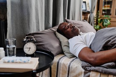 Aged African American man in white t-shirt sleeping under gray cover on comfortable double bed by small round table with alarm clock