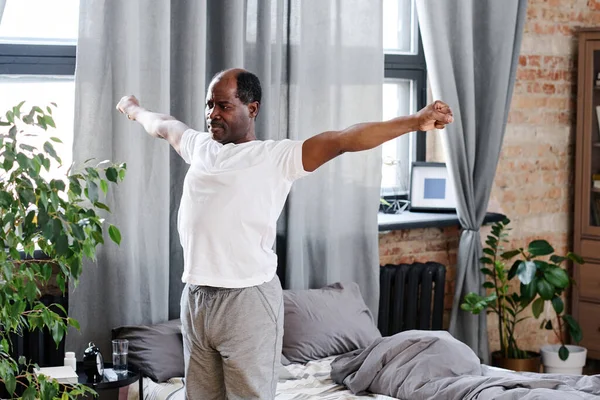 Active elderly black man with outstretched arms exercising in the morning while standing by his bed with grey pillows and blanket