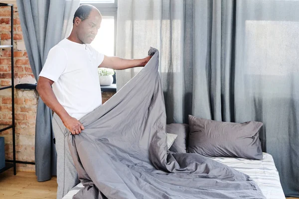 Contemporary African American old man in t-shirt making bed with grey blanket in the morning after sleep and physical exercises