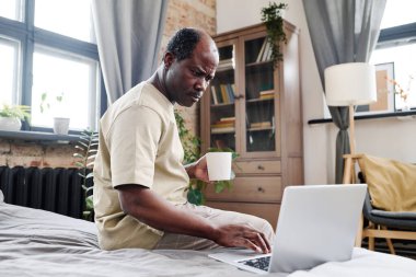 Serious retired African American man with cup of tea or coffee looking at laptop screen while sitting on bed and reading online news