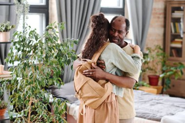 Cute schoolgirl with backpack embracing her African American grandfather while visiting him after school or on weekend