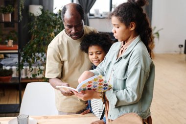 Contemporary senior black man helping his granddaughter with home assignment written in copybook while looking at her notes