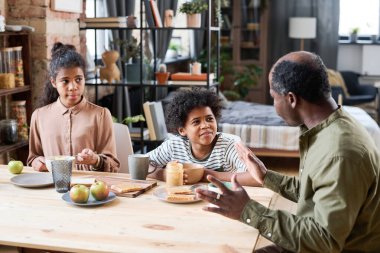 Contemporary retired black man talking to his grandchildren by table served with peanut butter, bread, apples and tea for breakfast