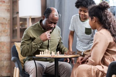 Pensive senior man looking at chessboard with white and brown wooden figures while playing with his two grandchildren at leisure