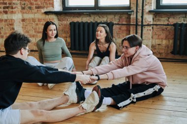 Two active teenage guys holding by hands while sitting in front of one another and touching by soles during vogue dancing repetition