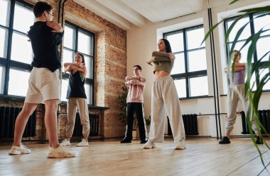 Group of teenage performers in activewear repeating vogue dancing exercises after male instructor while standing in front of him