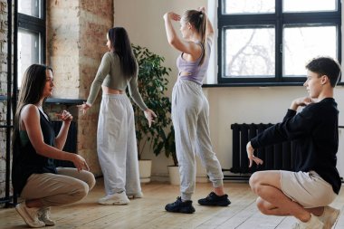 Young man showing new vogue dance movements to one of active teenage girls sitting in front of him and asking questions