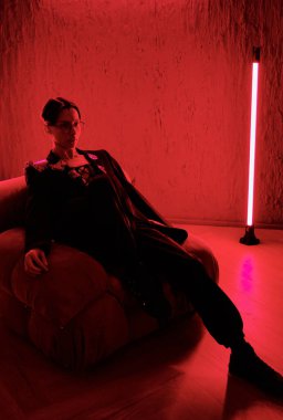 Young restful posh man or teenage guy in smart suit relaxing in armchair in night club in spacious stylish room lit with red light