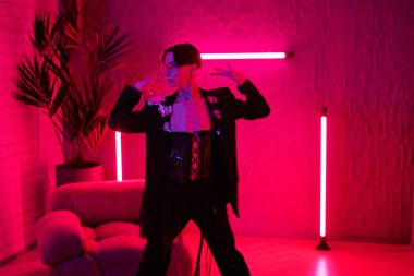 Contemporary young dancer in smart attire performing vogue dance in night club while standing in room or studio lit with pink neon light