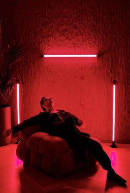 Contemporary young man in smart attire sitting in soft comfortable armchair in the room lit with red light in entertainment club