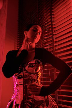 Contemporary teenage girl in posh stage dress posing in front of camera against venetian blinds in dance studio lit with red light