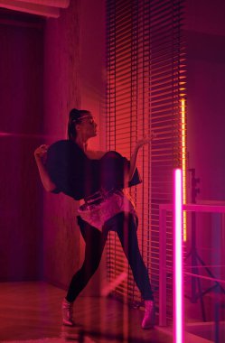 Young active female performer in futuristic attire dancing in studio lit with neon lamps while standing in front of venetian blinds