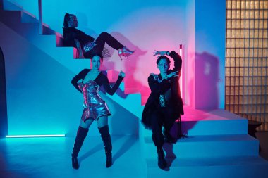 Young teenage couple of vogue dancers in posh attire performing against girl in glamorous apparel sitting on staircase in modern studio