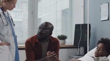 Tilting up and down of mature Caucasian female doctor coming and reassuring loving father of unconscious African American son who lying in bed in hospital ward at daytime