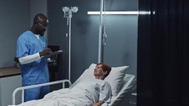 Medium long of young Black male nurse wearing blue scrubs taking notes, talking to female red-haired Caucasian patient lying in bed in hospital ward