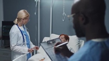Over-shoulder of blurred Black male nurse taking notes of blond mature doctor interviewing female senior Caucasian patient who lying in bed in hospital ward